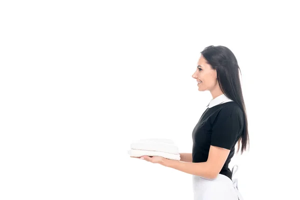 Vista laterale di cameriera sorridente in uniforme in possesso di asciugamani isolati su bianco — Foto stock