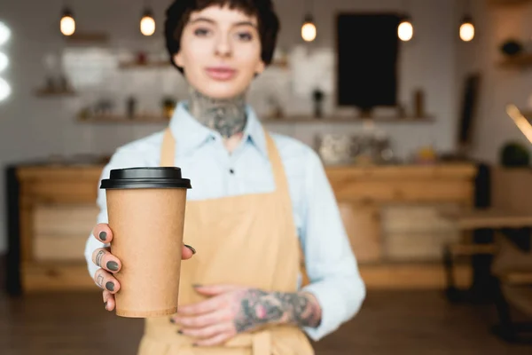 Foyer sélectif de serveuse attrayante dans tablier tenant tasse en papier et regardant la caméra — Photo de stock