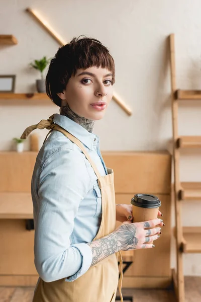 Jolie serveuse dans tablier tenant tasse jetable et regardant la caméra — Photo de stock