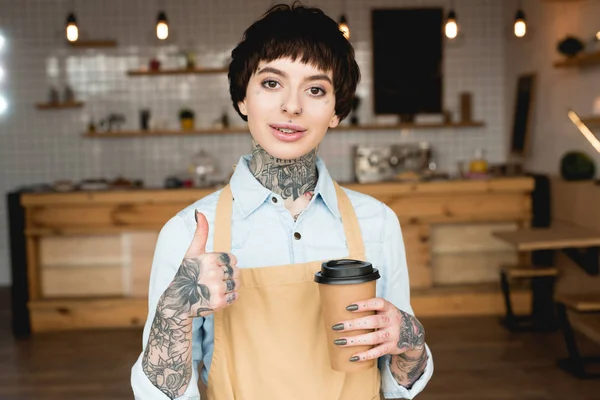 Camarera sonriente en delantal mostrando el pulgar hacia arriba y sosteniendo la taza de papel - foto de stock