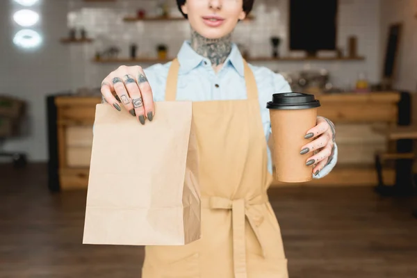 Vista parziale della cameriera tenendo sacchetto di carta e tazza usa e getta in mani tese — Foto stock