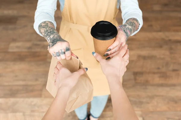Vista cortada de garçonete tatuado dando saco de papel e copo descartável para o cliente — Fotografia de Stock