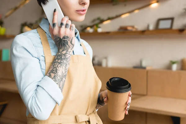 Abgeschnittene Ansicht einer Kellnerin, die mit Smartphone spricht und Einwegbecher in der Hand hält — Stockfoto
