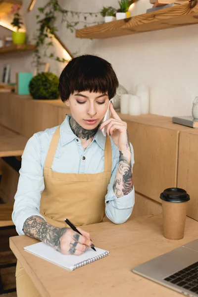 Концентрована офіціантка розмовляє на смартфоні і пише в блокноті — стокове фото
