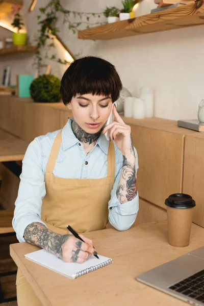 Serveuse concentrée parlant sur smartphone et écrivant dans un carnet — Photo de stock