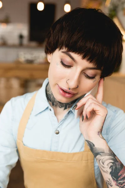 Atractiva camarera tatuada hablando en el teléfono inteligente y mirando hacia abajo - foto de stock