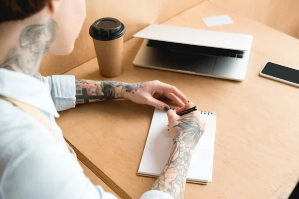 Overhead view of waitress writing in notebook while sitting at desk near laptop and paper cup — Stock Photo