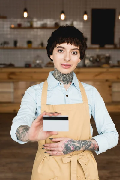 Camarera sonriente propietaria en delantal con tarjeta de crédito y mirando a la cámara - foto de stock