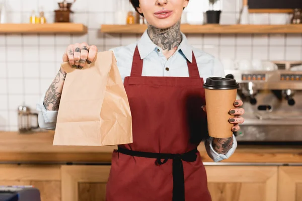 Partial view of barista in apron holding paper bag and disposable cup — Stock Photo