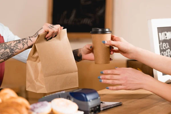 Partial view of barista holding paper bag and disposable cup near customer — Stock Photo