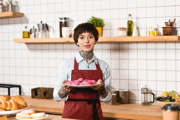 Hübsche Barista hält Gericht mit leckeren Keksen und schaut in die Kamera — Stockfoto