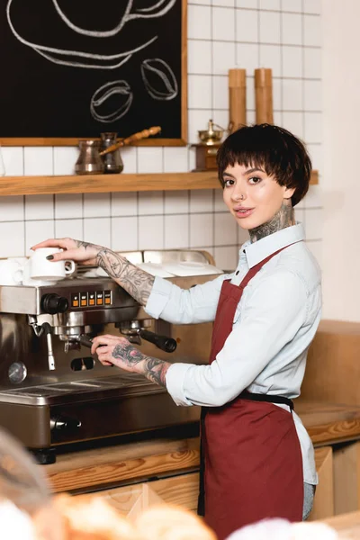 Selective focus of smiling barista preparing coffee on coffee machine and looking at camera — Stock Photo