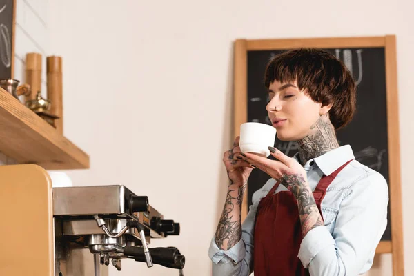 Bastante barista beber café con los ojos cerrados mientras está de pie en la máquina de café - foto de stock