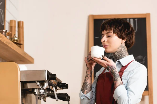 Grazioso barista bere caffè con gli occhi chiusi mentre in piedi alla macchina del caffè — Stock Photo