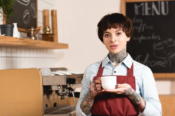 Bella barista in possesso di tazza di caffè mentre in piedi a macchina del caffè — Stock Photo