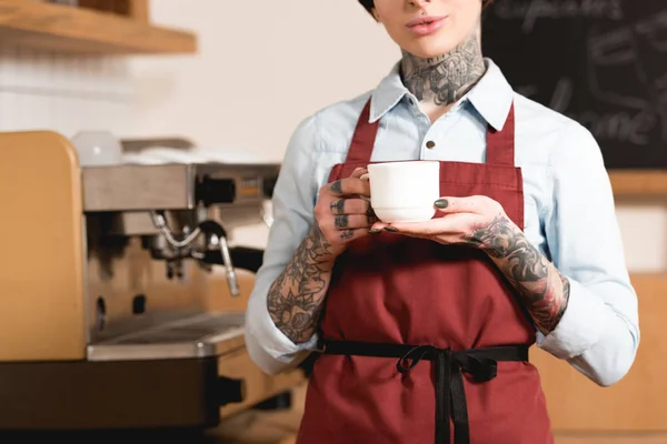 Частичный вид бариста в фартуке держа чашку кофе, стоя у кофеварки — стоковое фото