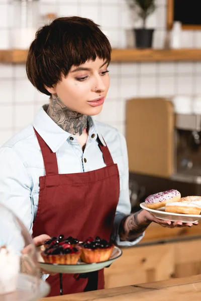 Hermosa barista sosteniendo platos con deliciosa pastelería en la cafetería - foto de stock