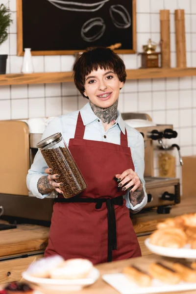 Selective focus of smiling barista holding jar with coffee grains and looking at camera — Stock Photo