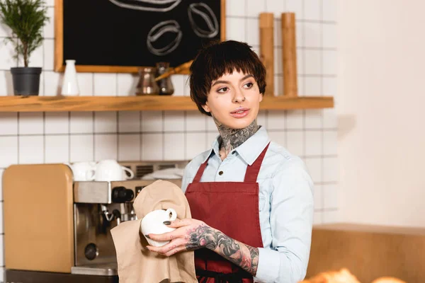 Selective focus of beautiful barista wiping cup while standing near espresso machine in cafe — Stock Photo