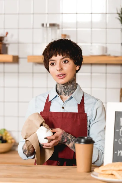 Attractive barista in apron wiping cup while standing near bar counter — Stock Photo