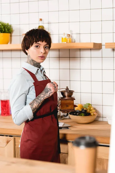 Selective focus of pretty barista in apron holding coffee mill and looking at camera — Stock Photo