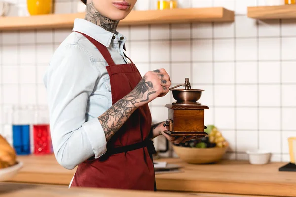 Частковий вид татуйованої бариста в фартусі, що тримає кавомолку — стокове фото