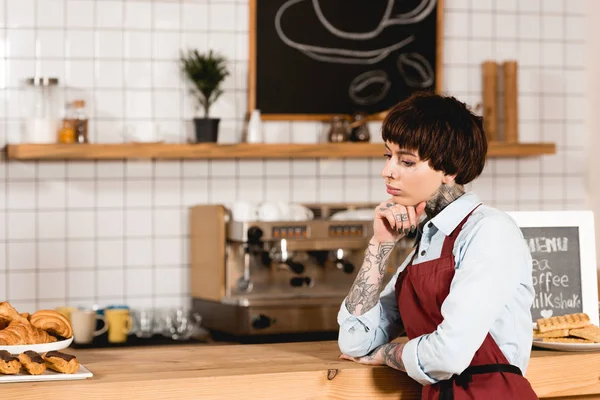 Barista pensieroso in grembiule in piedi vicino al bancone del bar in legno in caffetteria — Foto stock