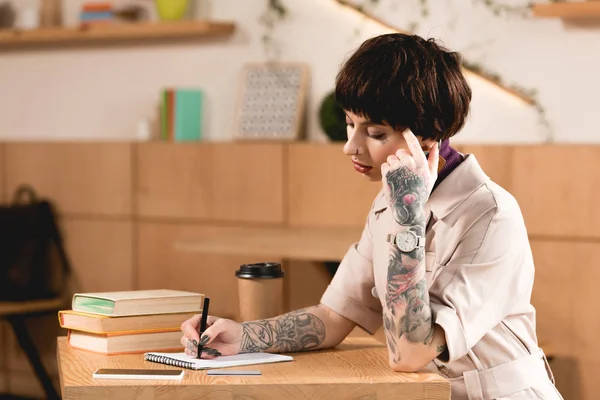 Красива бізнес-леді, що пише в блокноті, сидячи за столом біля книг і паперової чашки — стокове фото