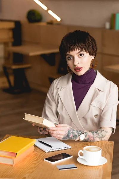 Atractiva y segura empresaria sentada en la cafetería y sosteniendo el libro - foto de stock