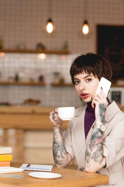 Attraente donna d'affari parlando su smartphone, tenendo in mano la tazza di caffè e guardando la fotocamera — Foto stock
