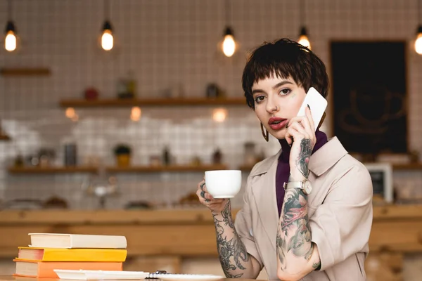 Atractiva mujer de negocios hablando en el teléfono inteligente, sosteniendo la taza de café y mirando a la cámara - foto de stock