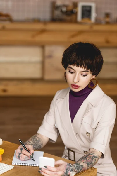 Attractive businesswoman writing in notebook and holding smartphone in coffee shop — Stock Photo