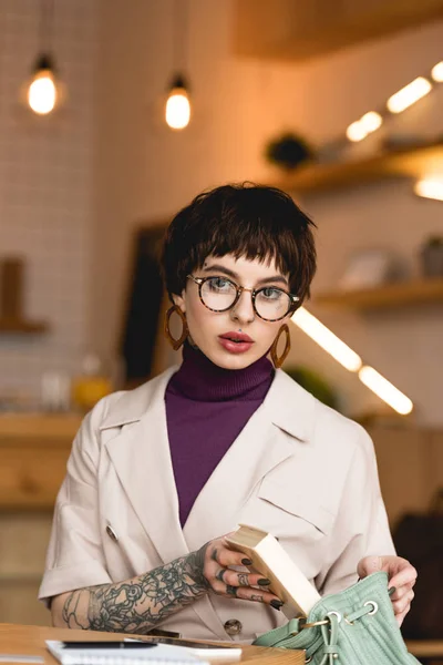 Stilvolle Geschäftsfrau mit Brille steckt Buch in Tasche und blickt in die Kamera — Stockfoto