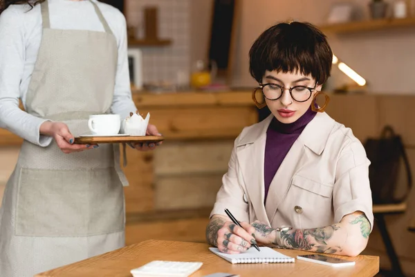 Vista parcial de la camarera sosteniendo bandeja cerca de la moderna mujer de negocios sentada a la mesa y escribiendo en un cuaderno - foto de stock