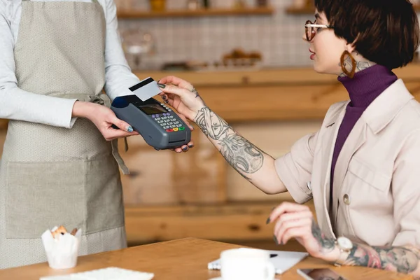 Vue partielle de la serveuse tenant terminal de paiement près d'une femme d'affaires avec carte de crédit — Photo de stock