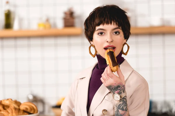 Moda, atractiva mujer de negocios degustación de deliciosa eclair y mirando a la cámara - foto de stock