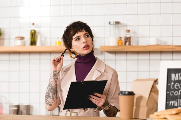 Thoughtful businesswoman holding clipboard while standing at bar counter in cafe — Stock Photo