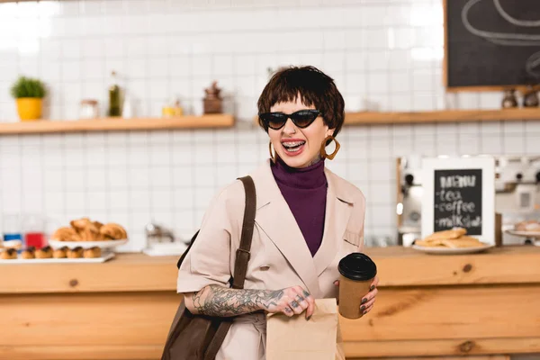 Sonriente, mujer de negocios de moda en gafas de sol sosteniendo taza desechable - foto de stock