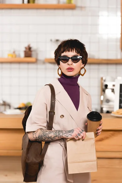 Fashionable businesswoman in sunglasses holding disposable cup and paper bag — Stock Photo