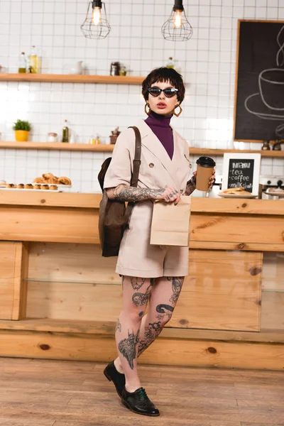 Hermosa, mujer de negocios de moda sosteniendo taza desechable y bolsa de papel en la cafetería - foto de stock
