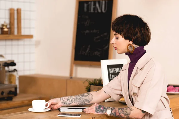 Attraktive Geschäftsfrau sitzt am Tresen neben Kaffeetasse, Notizbuch und Smartphone — Stockfoto