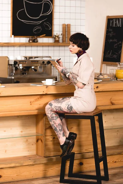 Mujer de negocios de moda y tatuada sentada en el mostrador del bar y escribiendo en un cuaderno - foto de stock