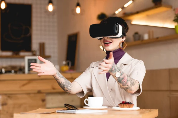 Улыбающаяся деловая женщина в наушниках виртуальной реальности сидит за столом с чашкой кофе и тортом в кафе — стоковое фото