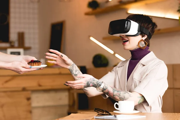 Smiling businesswoman in virtual reality headset taking saucer with cake from waitress — Stock Photo