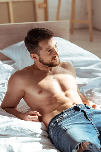 Hombre guapo y sin camisa acostado en la cama y mirando hacia otro lado en el dormitorio - foto de stock