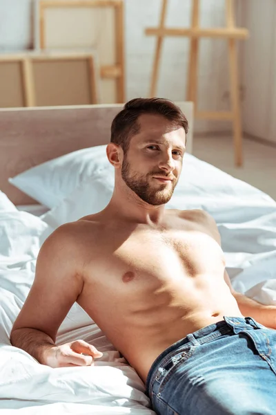 Bonito e sem camisa homem deitado na cama e olhando para a câmera no quarto — Fotografia de Stock