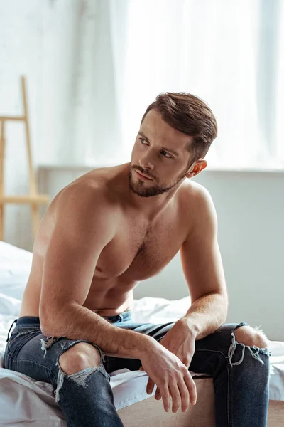 Hombre guapo y sin camisa sentado en la cama y mirando hacia otro lado en el dormitorio - foto de stock