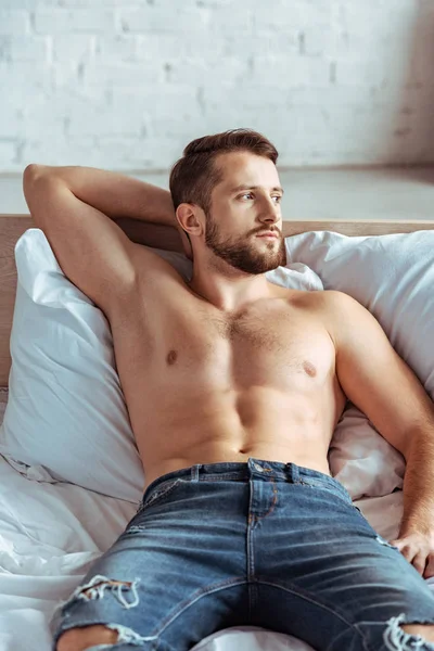 Good-looking and muscular man lying on bed and looking away in bedroom — Stock Photo