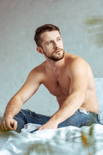 Homem bonito e musculoso sentado na cama e olhando para longe no quarto — Fotografia de Stock