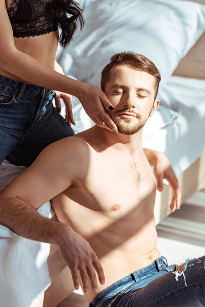 Mujer abrazando hombre guapo, musculoso y sin camisa con los ojos cerrados en el dormitorio - foto de stock